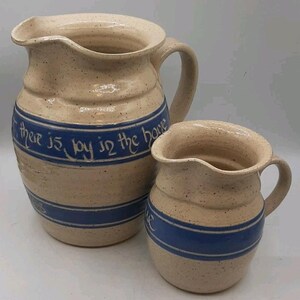 Puede incluir: Dos jarras de cerámica beige con rayas y asas azules. La jarra más grande tiene el texto azul "there is joy in the home". Las jarras tienen un acabado moteado y una apariencia rústica hecha a mano.