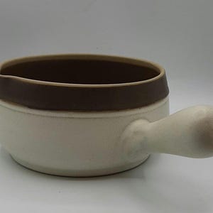 May include: A white ceramic sauce boat with a brown rim and a handle. The boat is designed for serving sauces or gravies.