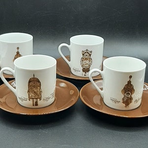 Könnte beinhalten: Vier weiße Keramik-Kaffeetassen mit braunen Untertassen. Jede Tasse zeigt eine andere, vintage-inspirierte Illustration eines Uhrturms. Die Tassen sind auf einer braunen Oberfläche angeordnet.