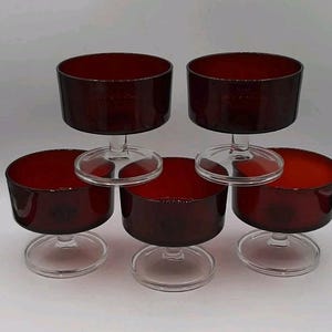 May include: A set of five matching dessert bowls made of deep red glass. Each bowl features a wide, cylindrical basin supported by a short, clear glass stem and a circular base. The bowls are arranged in a group with two in the back and three in the front.