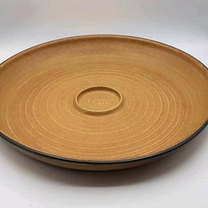 May include: A large, round serving platter with a light brown surface and a dark rim. The center features a smaller, circular indentation. The platter has concentric circle design and is suitable for serving food.