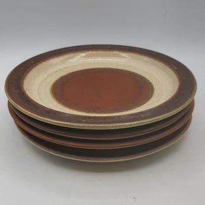 Puede incluir: Una pila de cuatro platos de cerámica. Cada plato presenta un borde marrón, una banda beige y un centro marrón rojizo. Los platos están apilados, mostrando su diseño en capas.