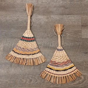 May include: Two hand-woven straw fans with long handles. The fan heads are triangular, featuring woven patterns with bands of colour including red, green, yellow, and black. The handles are wrapped with twine.