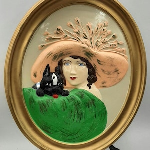 May include: Oval framed relief sculpture depicting a woman in a large peach hat with a black dog. The dog rests on a green cushion. The frame is gold-coloured, and the background is off-white.