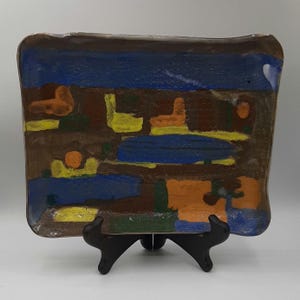 May include: A rectangular ceramic plate with an abstract design. The plate features a brown border and a central design with blue, yellow, and orange shapes. It is displayed on a black stand.