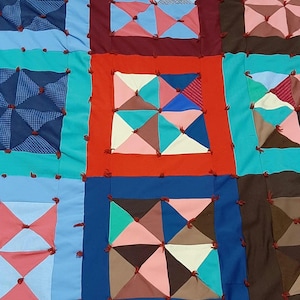 May include: A patchwork quilt featuring a range of colours and patterns, including blue, red, green, brown, and pink. The quilt is composed of squares, each with a unique design. The quilt is tied together with red string.