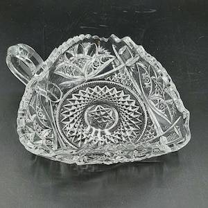 May include: Clear, triangular-shaped glass dish with a handle. The dish features intricate cut glass patterns, including stars, geometric shapes, and floral designs. The handle is curved and integrated into the design. The dish is likely for serving.