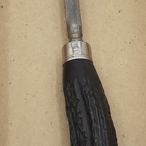 Puede incluir: Una lima met&aacute;lica con una lima plateada estriada y un mango negro texturizado. El mango tiene una forma curvada y un collar plateado. Esta herramienta probablemente se utiliza para dar forma o alisar materiales.