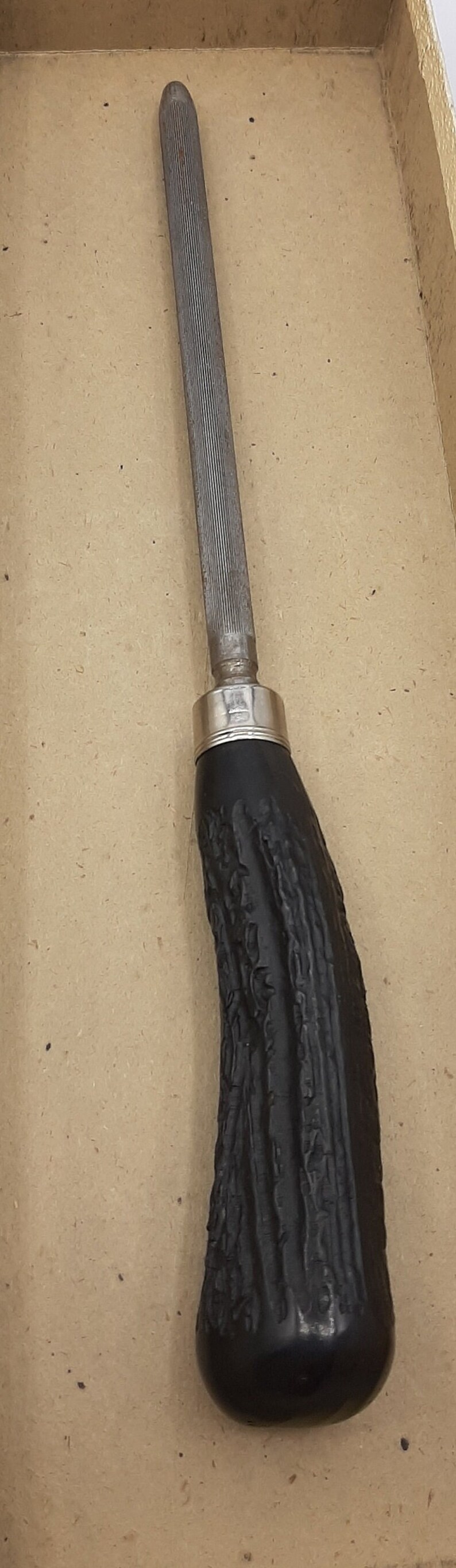 Puede incluir: Una lima met&aacute;lica con una lima plateada estriada y un mango negro texturizado. El mango tiene una forma curvada y un collar plateado. Esta herramienta probablemente se utiliza para dar forma o alisar materiales.