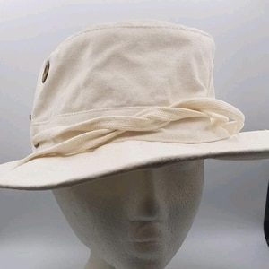 May include: A cream-colored bucket hat featuring a wide brim and a braided fabric band encircling the crown. The hat includes metallic eyelets on the sides for ventilation, constructed from a solid, light-colored textile material.