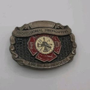 Może przedstawiać: Owalna metalowa klamra do paska w kolorze ciemnoszarym z centralnym emblemem krzyża maltańskiego w kolorze czerwonym i kremowym. Napis głosi PROFESSIONAL FIREFIGHTERS u góry i AMERICA'S HEROES u dołu, z literami F i D po bokach.