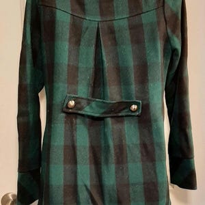 May include: A green and black checked coat with a notched collar and long sleeves. The back has a decorative flap with two gold-coloured buttons. The coat has a tailored fit and a classic design.