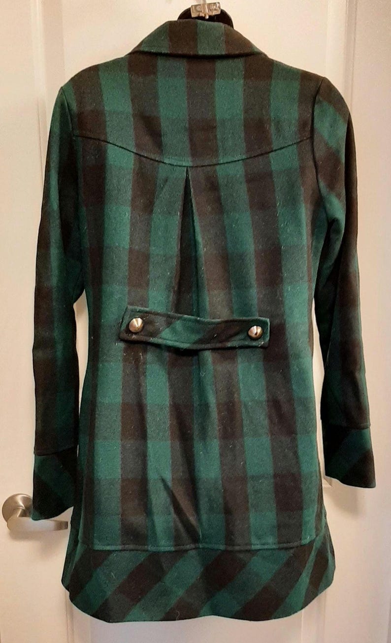 May include: A green and black checked coat with a notched collar and long sleeves. The back has a decorative flap with two gold-coloured buttons. The coat has a tailored fit and a classic design.