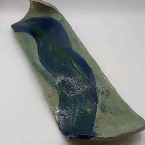 May include: Handmade ceramic serving dish. The rectangular dish features a wavy design with a raised edge. The color palette includes shades of blue and green, creating a unique, artistic aesthetic. The dish is approximately 12 inches long.