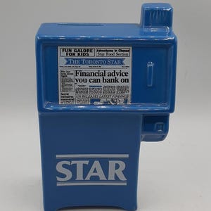 May include: A blue ceramic bank shaped like a newspaper vending machine. The front features the word "STAR" in white block letters. The top has a slot for inserting money. The side has a small rectangular opening. The front has text that reads "THE TORONTO STAR".