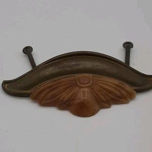 May include: Antique brass-toned drawer pull with a carved, light brown, floral-like design. The pull has two screws for mounting. The curved handle is a darker brass color.
