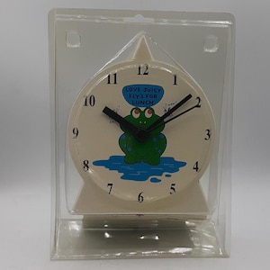 May include: A cream-colored novelty clock featuring a cartoon frog with the text "LOVE JUICY FLYS FOR LUNCH". The clock has black hands and numbers, and is encased in a clear plastic package.