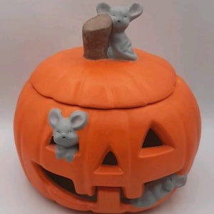 May include: A bright orange ceramic pumpkin jar with a removable lid. The pumpkin features a carved jack-o'-lantern face with triangular eyes and a wide mouth. Three small, light grey sculpted mice are attached to the exterior surface.
