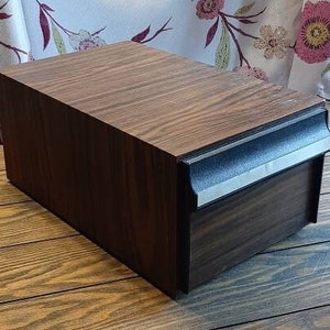 Può includere: Una scatola portaoggetti rettangolare in legno con finitura marrone scuro. La scatola ha un cassetto con finiture nere e argentate. La venatura del legno è visibile sulla parte superiore e sui lati della scatola. La scatola è appoggiata su una superficie di legno.