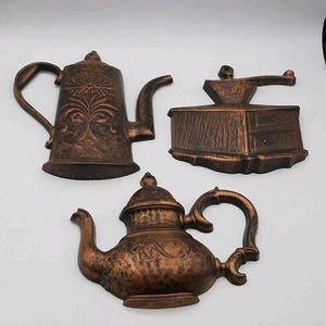 Puede incluir: Tres placas de pared de color cobre con diseños en relieve de una cafetera alta con motivo floral, un molinillo de café manual con manivela y una tetera redondeada con un patrón de volutas decorativo en el lateral del cuerpo.