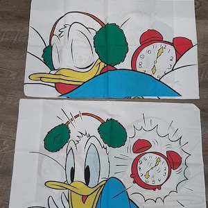 Könnte beinhalten: Zwei Kissenbezüge mit einem Comic-Enten-Motiv, das Ohrenschützer trägt und einen Wecker zeigt. Die Ente schläft im ersten Kissenbezug und wird im zweiten Kissenbezug vom Wecker geweckt.