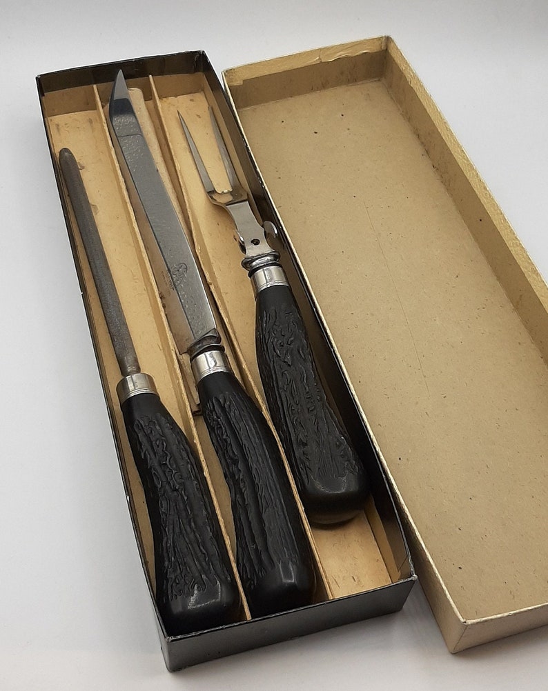Puede incluir: Un juego de trinchar vintage en una caja de cart&oacute;n. El juego incluye un cuchillo para trinchar, una chaira y un tenedor para trinchar. Los mangos son negros con un dise&ntilde;o texturizado, y las hojas y el tenedor son plateados. La caja est&aacute; abierta.