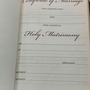 May include: Open book with a Certificate of Marriage page. The text reads "Certificate of Marriage", "Holy Matrimony", and includes lines for names, dates, and witnesses. The page is cream-colored with brown text.