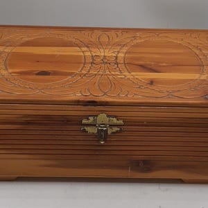 Puede incluir: Una caja de madera rectangular con tapa con bisagras. La tapa presenta diseños florales tallados y la caja tiene un cierre de color latón. La madera tiene un tono cálido y natural.