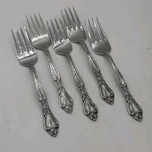 May include: Six silver forks with ornate handles are arranged diagonally. The forks have tines and decorative detailing on the handles. The silverware is likely for formal dining and adds a touch of elegance to any table setting.
