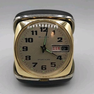 May include: A vintage Bulova Day & Date Alarm clock. The clock has a gold-tone face with black numerals and hands. The day and date are displayed in a small window. The clock is encased in a black and gold folding case.