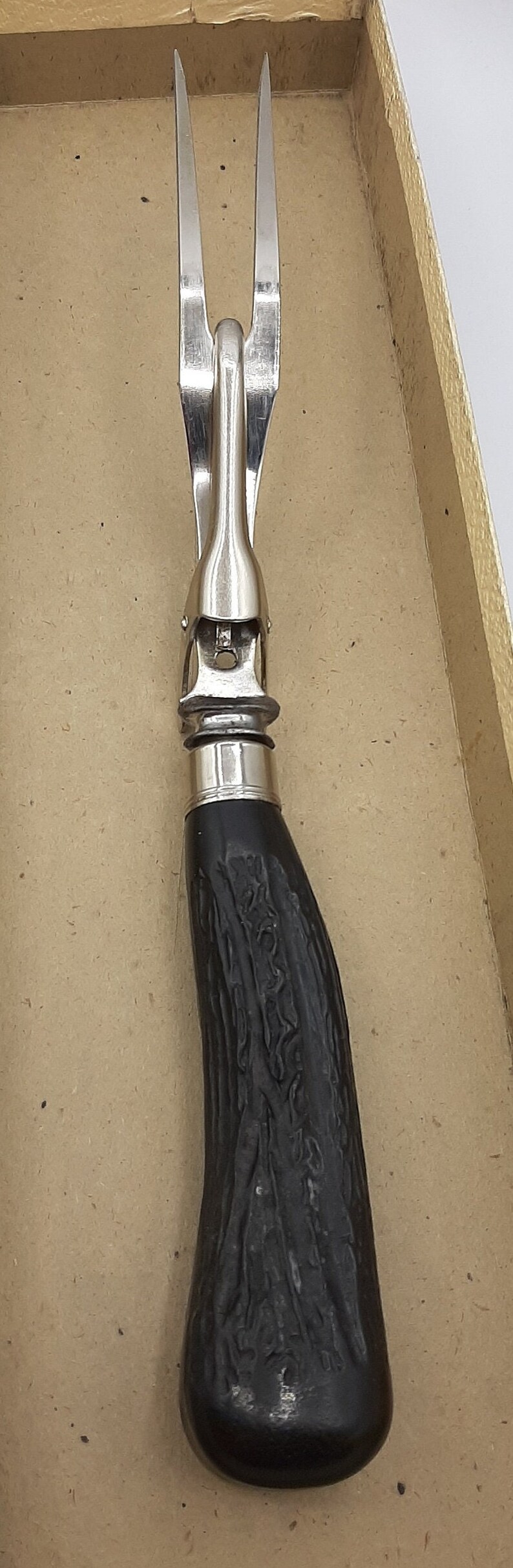 Puede incluir: Un tenedor para trinchar vintage con cabeza de acero inoxidable y mango oscuro y texturizado. El tenedor tiene dos p&uacute;as y una virola decorativa. El mango parece ser de madera tallada o similar.