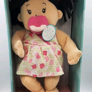 May include: A plush Baby Stella doll with black yarn hair in pigtails, wearing a floral dress. The doll has a pink pacifier and is packaged in a box. The box has the Baby Stella logo.