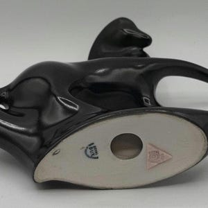 May include: A glossy black ceramic figurine of a stylized animal, possibly a horse or a deer, with a long, curved neck and a sleek, modern design. The underside features a light-colored base with a circular opening and a pink triangle emblem.