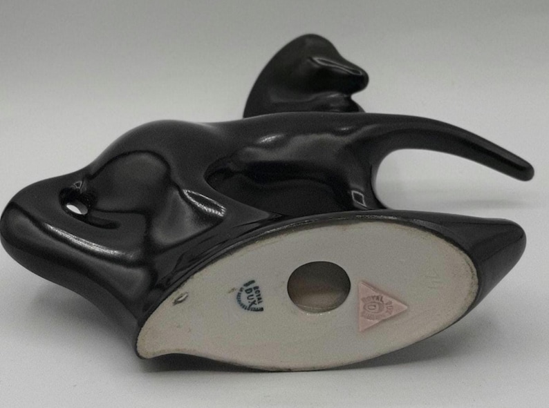 May include: A glossy black ceramic figurine of a stylized animal, possibly a horse or a deer, with a long, curved neck and a sleek, modern design. The underside features a light-colored base with a circular opening and a pink triangle emblem.