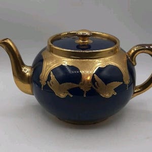 May include: A navy blue teapot with gold detailing. The teapot has a rounded shape, a curved spout, and a looped handle. Gold embellishments and bird designs decorate the surface. The lid is navy blue with a gold knob.