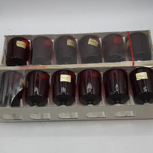 May include: A set of twelve dark red drinking glasses in a cardboard box. The glasses are cylindrical with a slightly flared rim and a thick base. Each glass has a small, rectangular sticker. The box is white with a clear plastic window.