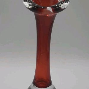 May include: A tall, slender bud vase crafted from clear glass with a deep red interior core. The vase features a wide, flared rim and a thick, rounded base containing visible air bubbles, creating a distinct contrast between the red and clear glass.