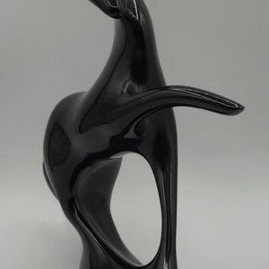 May include: A glossy black abstract sculpture with a smooth, curved design. The sculpture has a flowing form with an open space in the center and an extended arm-like element. The artwork is set against a plain background.
