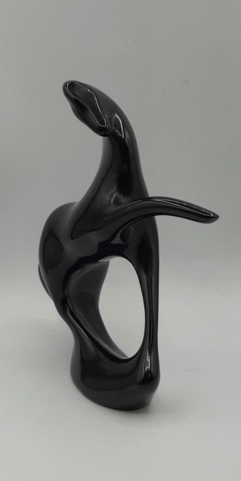 May include: A glossy black abstract sculpture with a smooth, curved design. The sculpture has a flowing form with an open space in the center and an extended arm-like element. The artwork is set against a plain background.