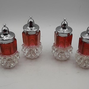 May include: Four vintage salt and pepper shakers. Each shaker has a faceted clear glass base, a red, faceted body, and a silver-colored metal lid. The shakers are arranged in a row against a white background.