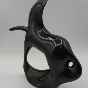 May include: A glossy black decorative mask with a curved, horn-like top and a small, protruding side element. The mask has a large, oval-shaped eye opening and a small, circular hole near the bottom.