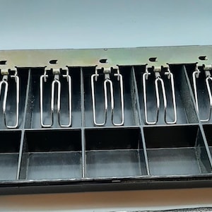 May include: An open black cash register drawer with five compartments, each with a metal spring clip. The drawer's interior is visible. The design is functional and utilitarian.
