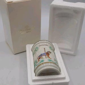 May include: A cylindrical porcelain spice jar labeled Coriander, featuring a carousel tiger illustration with gold trim. The jar is nestled in white styrofoam packaging next to its matching off-white cardboard box with gold text.
