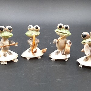 Puede incluir: Cuatro pequeñas figuritas de rana hechas de conchas marinas, cada una tocando un instrumento diferente. Las ranas son verdes con grandes ojos negros y llevan sombreros de conchas. Los instrumentos incluyen una flauta, un saxofón, un tambor y platillos.