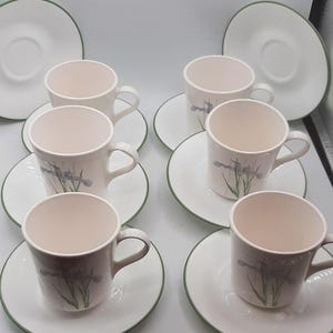 Pode incluir: Um conjunto de seis chávenas e pires a condizer. As chávenas e os pires são brancos com uma borda verde. Cada chávena tem um desenho floral em tons de roxo e verde. As chávenas estão dispostas em pires brancos.