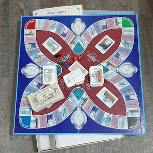 May include: A vintage board game, "Yuppie Game," with a blue and red game board. The board features various spaces, cards, dice, and play money. The game is made by Waddingtons.