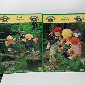 May include: Two Cabbage Patch Kids jigsaw puzzles. Each puzzle depicts Cabbage Patch Kids dolls in a forest scene. One shows dolls by a stream, the other by a bird's nest. The puzzles are labelled "Puzzle Casse-tête" and "AGE +7".