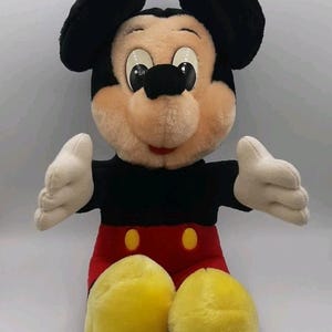 May include: A plush Mickey Mouse doll, featuring black ears, a tan face, and a red and black outfit. The doll has white gloves and yellow shoes. The iconic character is in a sitting position with arms outstretched.