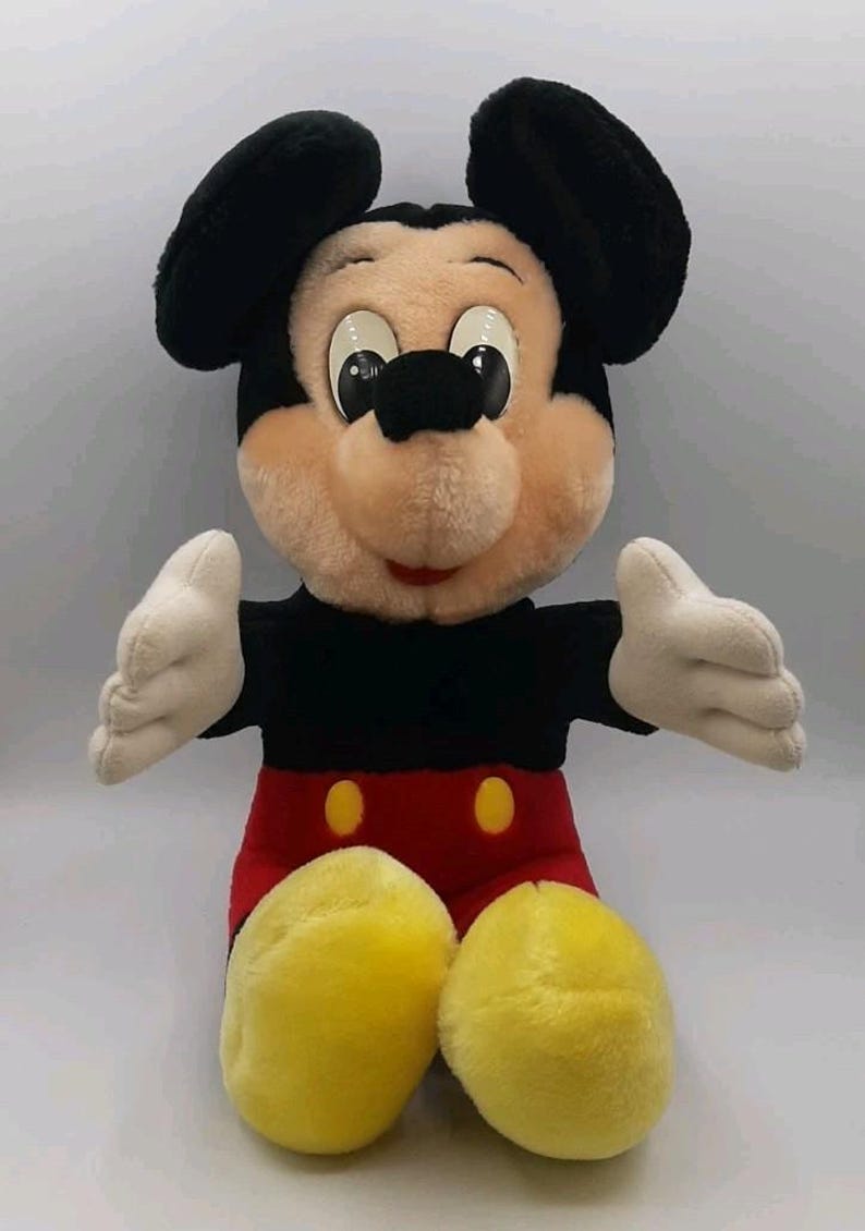 May include: A plush Mickey Mouse doll, featuring black ears, a tan face, and a red and black outfit. The doll has white gloves and yellow shoes. The iconic character is in a sitting position with arms outstretched.