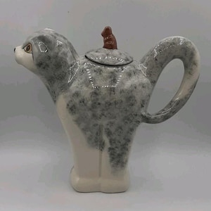 Puede incluir: Una tetera de cerámica con forma de gato con pelaje gris y blanco moteado. La tetera presenta una cabeza de gato con ojos marrones, un asa curva que forma la cola y una tapa extraíble rematada con una pequeña figura de color marrón.